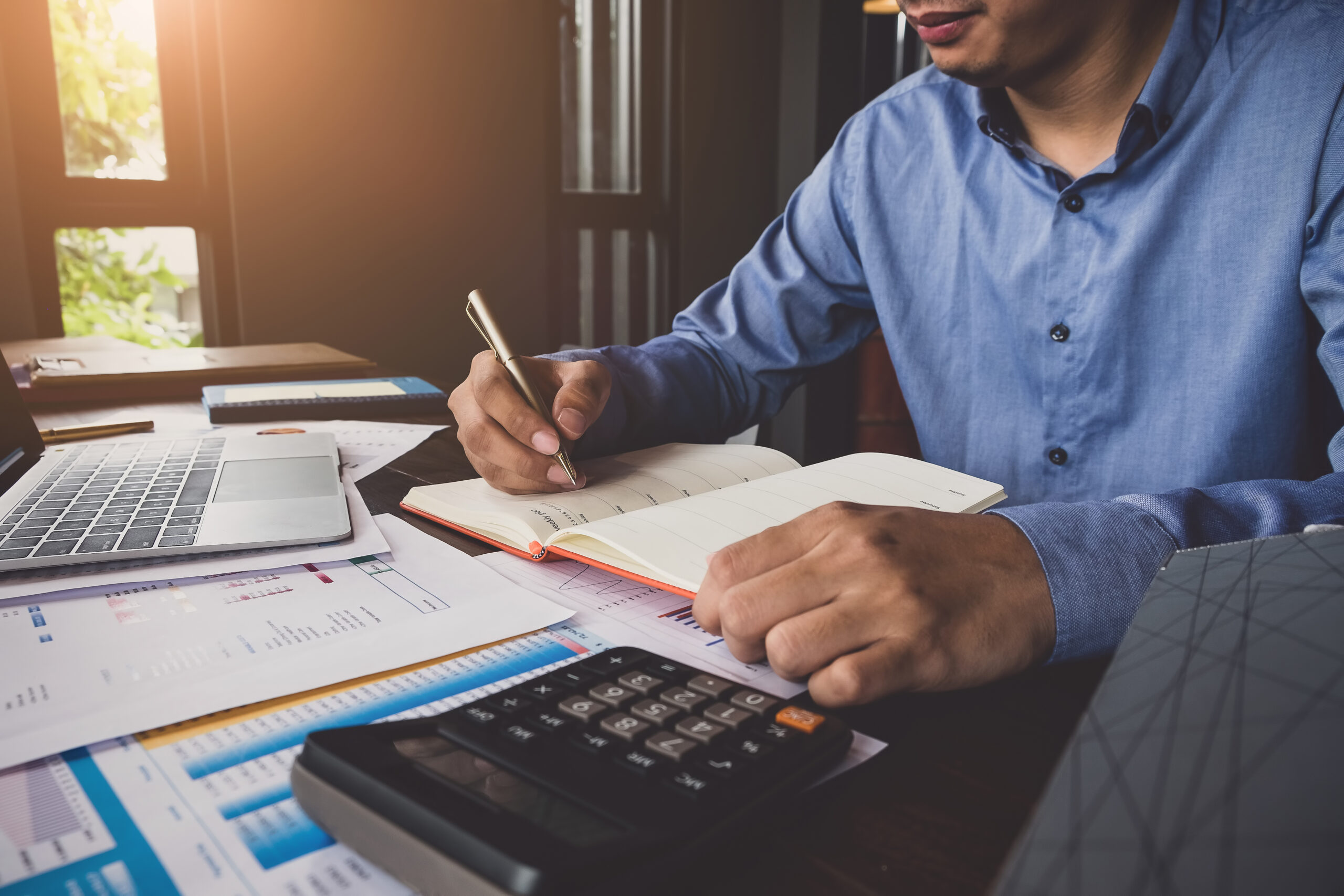 Man in blue shirt writes in a notebook at a desk with documents, a calculator, and a laptop, working in a well-lit office.